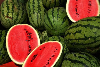 Many whole watermelons are stacked on top of one another, and some are cut open to show the red flesh and black seeds inside